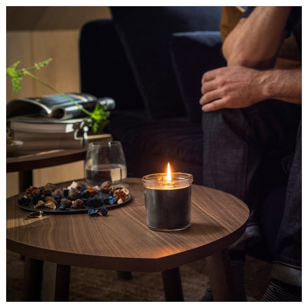 A person sits by a table with a lit FRUKTSKOG candle, clear glass, emitting a warm glow. Beside it, a plate with mixed nuts and a glass of water.