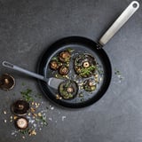 Top view of black non-stick frying pan with mushrooms, herbs, and oil. Silver spatula beside. Gray background with scattered mushrooms and seasoning.