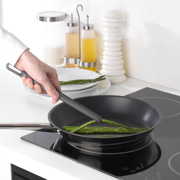 In a person is cooking asparagus with a black FULLÄNDAD cooking tweezers in a skillet on a modern black stovetop.