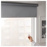 Hand raising remote to control gray roller blind in window.