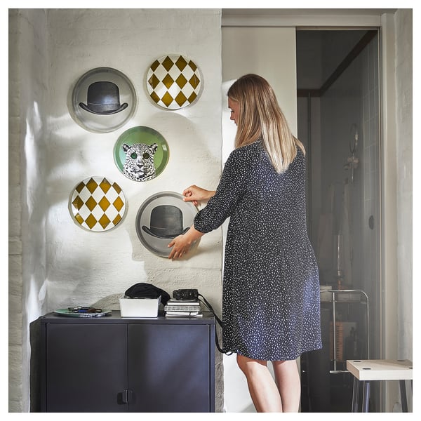 Woman adjusts hat wall decor; harlequin, leopard, and hat plates on gray wall, with black bookshelf.