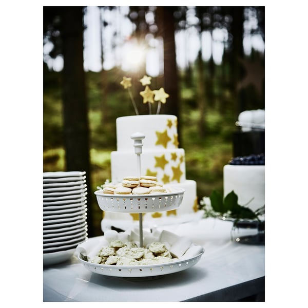 White, two-tiered serving stand with gold accents displays cookies and a cake, positioned on an outdoor table surrounded by greenery.