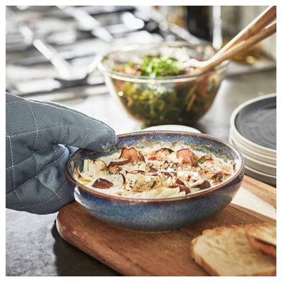 A hand grabs a blue glazed ceramic dish filled with baked food, next to a salad bowl and bread on a wooden board.
