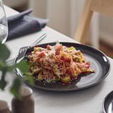 Plate of pasta with cherry tomatoes and grated cheese on a dark plate with fork, set on dining table.