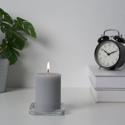 Lighted gray candle in glass holder near plant and clock.