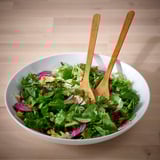 A large bowl of fresh green salad with wooden tongs on a wooden table.