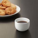 A small, white ceramic cup with a geometric design, containing coffee, sits on a table next to a plate of assorted cookies.