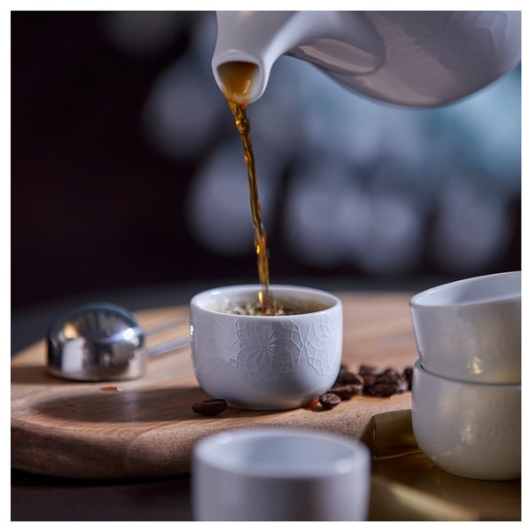 A white feldspar porcelain cup with a geometric pattern being filled from a pitcher. Surrounded by more cups and coffee beans.