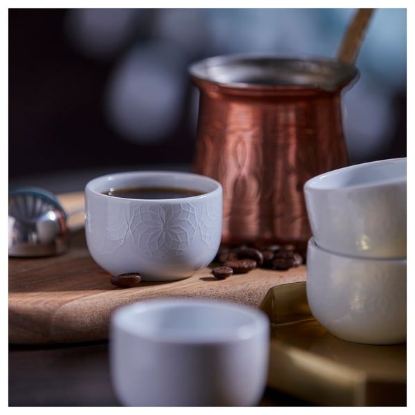 Small white porcelain cups with etched design, ideal for coffee or tea, sit on a rustic wooden table with a copper kettle and scattered coffee beans.