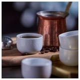 Small white porcelain cups with etched design, ideal for coffee or tea, sit on a rustic wooden table with a copper kettle and scattered coffee beans.