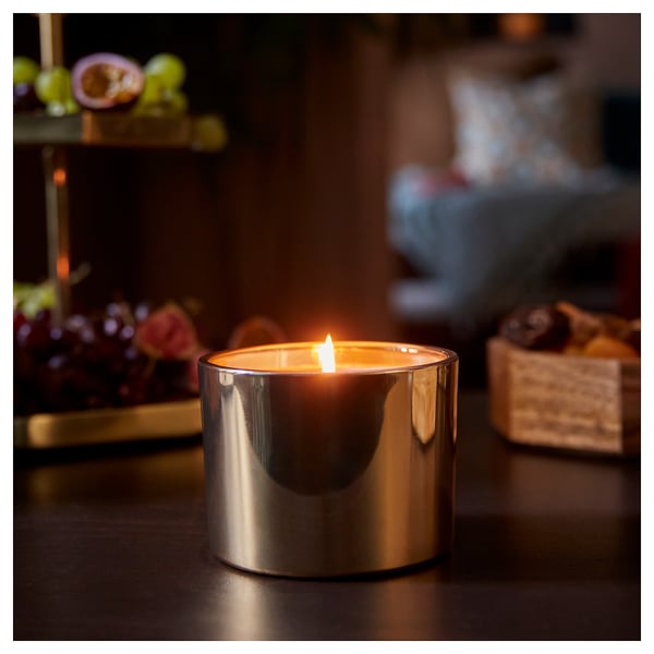 A lit GOKVÄLLÅ candle in a silver glass holder on a wooden table. The flame casts a warm glow, surrounded by dark, blurred decorations.