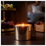 A lit GOKVÄLLÅ candle in a silver glass holder on a wooden table. The flame casts a warm glow, surrounded by dark, blurred decorations.
