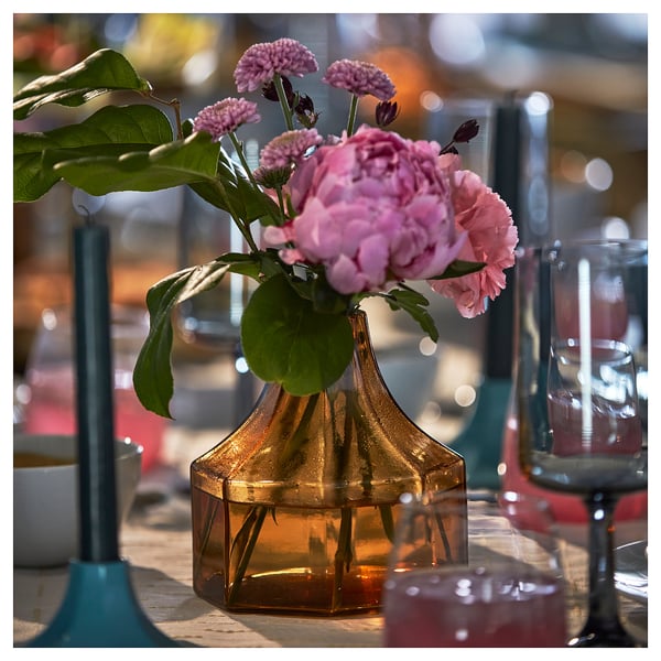 A yellow octagonal glass vase with pink flowers on a wooden table. Part of the GOKVÄLLÅ collection.