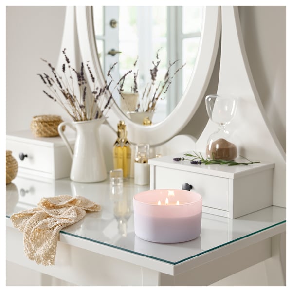 A cozy vanity setup with a lit GOLIATTUJA lavender scented candle, featuring a glass container and tin lid, beside a white jug, vase, and mirror.