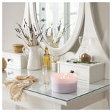 A cozy vanity setup with a lit GOLIATTUJA lavender scented candle, featuring a glass container and tin lid, beside a white jug, vase, and mirror.
