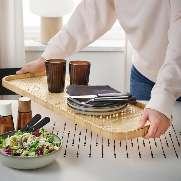 Person carrying bamboo tray with plates, cutlery, and glasses, and placing salad bowl on table.