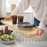 Person carrying bamboo tray with plates, cutlery, and glasses, and placing salad bowl on table.