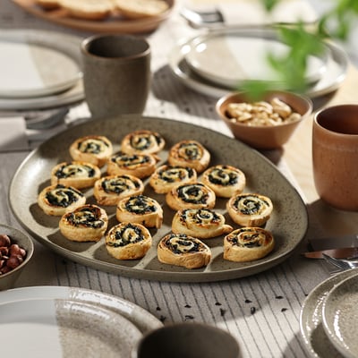 Large grey GRÅRÄV stoneware plate holds 16 pastry snacks, surrounded by smaller plates, cups, and bowls on a dining table.