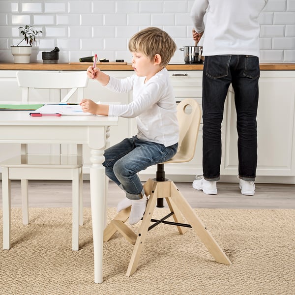 A young boy sits in an adjustable white highchair, drawing at a table; an adult stands nearby.