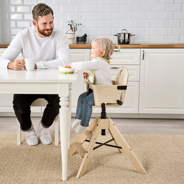 Man in white sweater drinks from mug, smiling at toddler in high chair. Chair has adjustable height and safety belt.