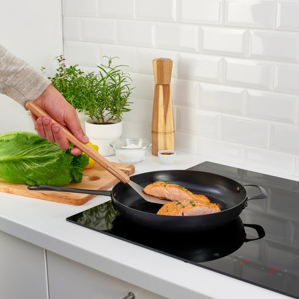 Person grilling fish in kitchen using GRILLTIDER tongs. Modern, clean kitchen with herbs and cooktop.