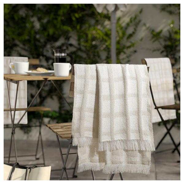 A cozy, checkered throw blanket with fringes hangs on a chair, ideal for outdoor use or chilly evenings, made from recycled polyester.