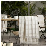 A cozy, checkered throw blanket with fringes hangs on a chair, ideal for outdoor use or chilly evenings, made from recycled polyester.