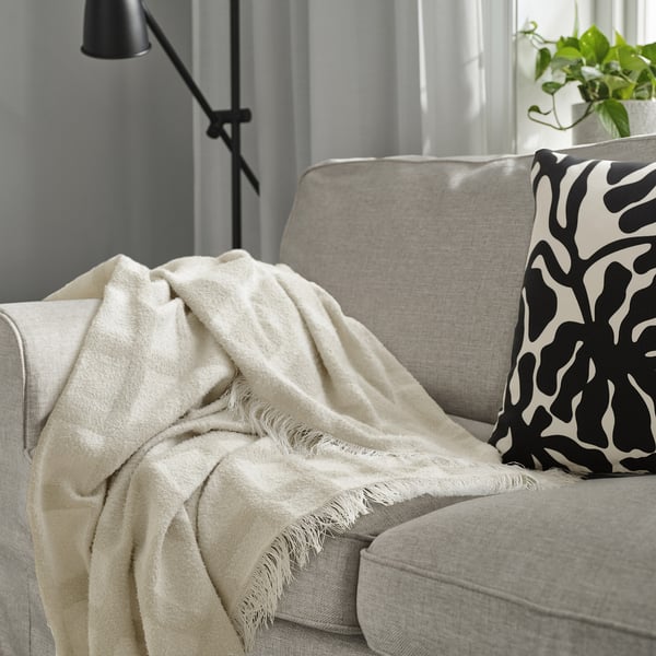 Beige GRÖNKNAVEL throw on grey sofa, with fringes and recycled polyester. Near black/white pillow and green plant.