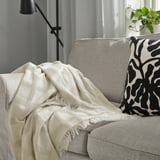 Beige GRÖNKNAVEL throw on grey sofa, with fringes and recycled polyester. Near black/white pillow and green plant.