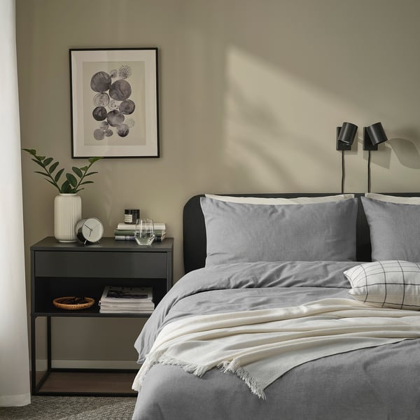 Bedroom scene with grey GROVSNÄRJA flannel bedding; twin black nightstands, one holding clock and plant, the other with books and lamp.
