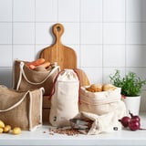 Textile bags in beige holding food, cotton, ideal for baked goods & veggies. Wooden board & greenery visible.