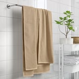 Beige GULVIAL cotton towel hangs on rack, surrounded by white bathroom decor, including a potted plant and neatly stacked towels on a white shelf.