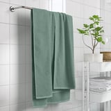 Green GULVIAL towel hangs on rack near white tiles, smaller than stacked towels, beside small potted plant.