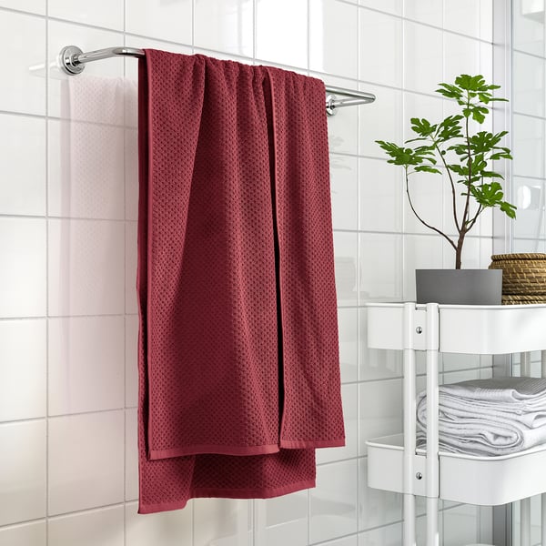 Red GULVIAL towel hangs on bar, near white shelves with folded towels, plant & basket, against tiled wall.