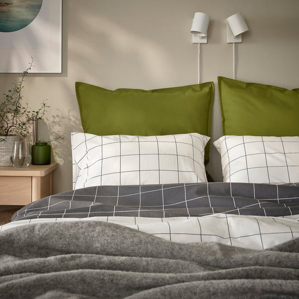 A bed with green GURLI pillowcases, white grid-patterned sheets, and grey blanket. Two wall lamps above, a plant beside.