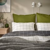 A bed with green GURLI pillowcases, white grid-patterned sheets, and grey blanket. Two wall lamps above, a plant beside.