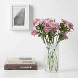 Pink flowers in HÄCKOXEL glass vase next to frames and books.