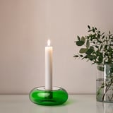 Green glass candlestick holder with lit white candle, beside vase with greenery.