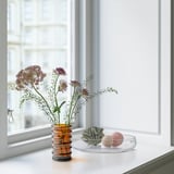 Vase on windowsill holds purple flowers, using recycled glass. Beside it, a dish with spheres and a folded item.
