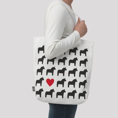 A person carrying a white tote bag with black horse patterns. A red heart is near the bottom.