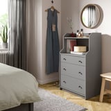 Modern bedroom with gray HAUGA chest, round mirror, blue dress, plant, wooden stool, bed with white blanket, gray curtains, wooden floor.