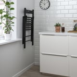 Small modern kitchen with white cabinets, a hanging black foldable chair, and a clock on white tiles. A plant on the windowsill adds greenery.