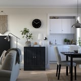 Modern living room with grey sofa, wooden cabinet, plants, and kitchen in background.