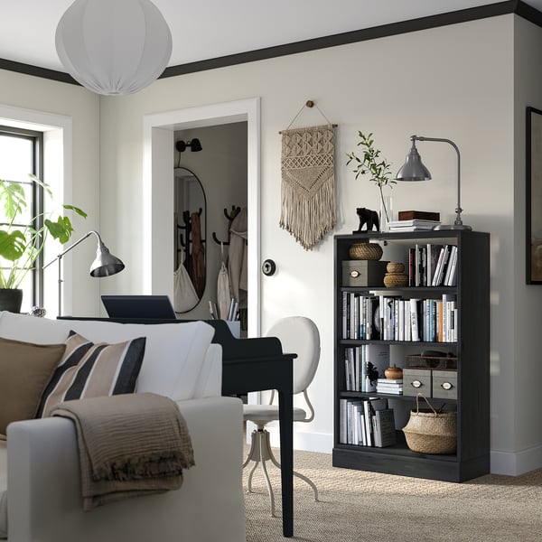 A stylish home office a white sofa, a black desk with a laptop, and a dark wooden bookshelf filled with books and decorated with a lamp and plants.