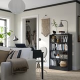 A stylish home office a white sofa, a black desk with a laptop, and a dark wooden bookshelf filled with books and decorated with a lamp and plants.