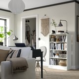 Bright living space with modern decor: white sofa, gray chair, wooden bookshelf with books, plants, and a black lamp.