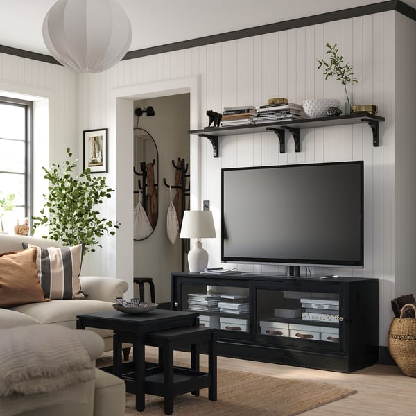 Modern living room with black tv stand, flat screen, beige sofa, pillows, coffee table, lamp, plants, and wall shelves with books.