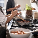 Two people cooking together on a gas stove, one stirring, the other prepping.