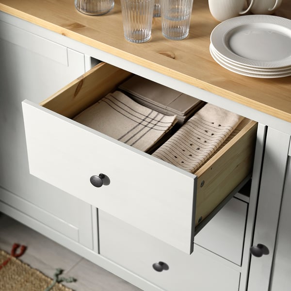 Open white HEMNES sideboard drawer with stacked napkins. Wood countertop with glasses and plates.