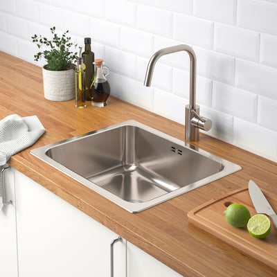 Stainless steel kitchen sink with faucet on wooden counter. Limes and knife on cutting board, bottles and plant nearby.